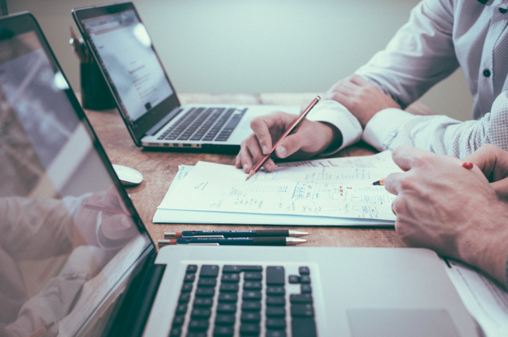 Photo of two men's hands writing on a piece of paper with laptops in the background