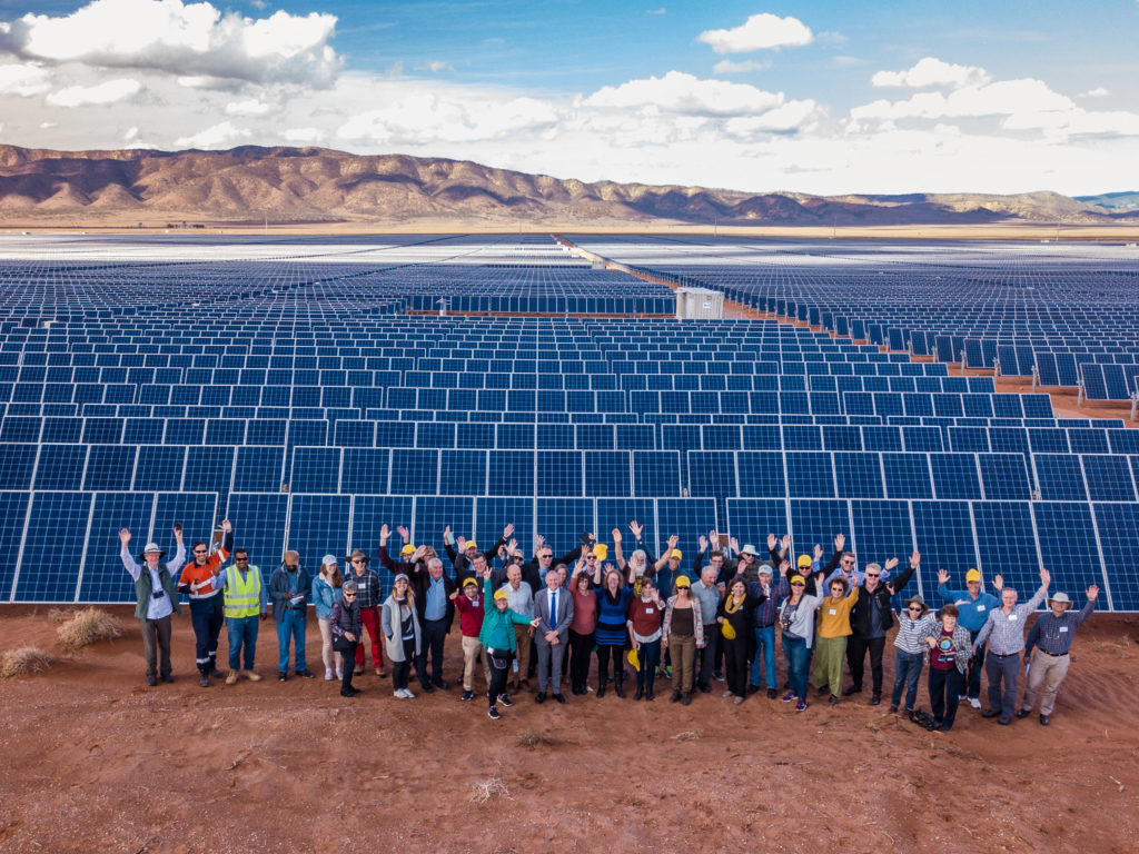 A photograph of people standing in front of a solar farm and waving