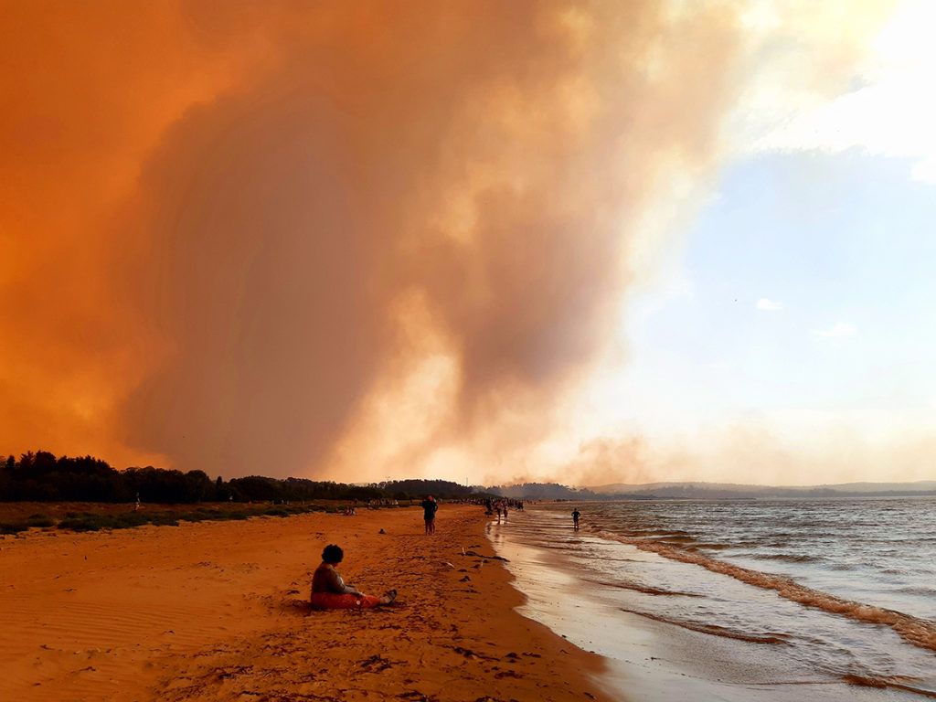 An image of a woman sitting on a beach, with orange sky from the smoke from bushfires.