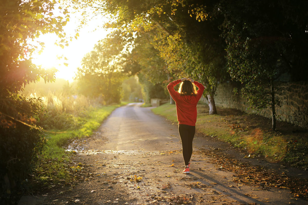 A woman with her hands above her head walking down a path with the sun streaming through.