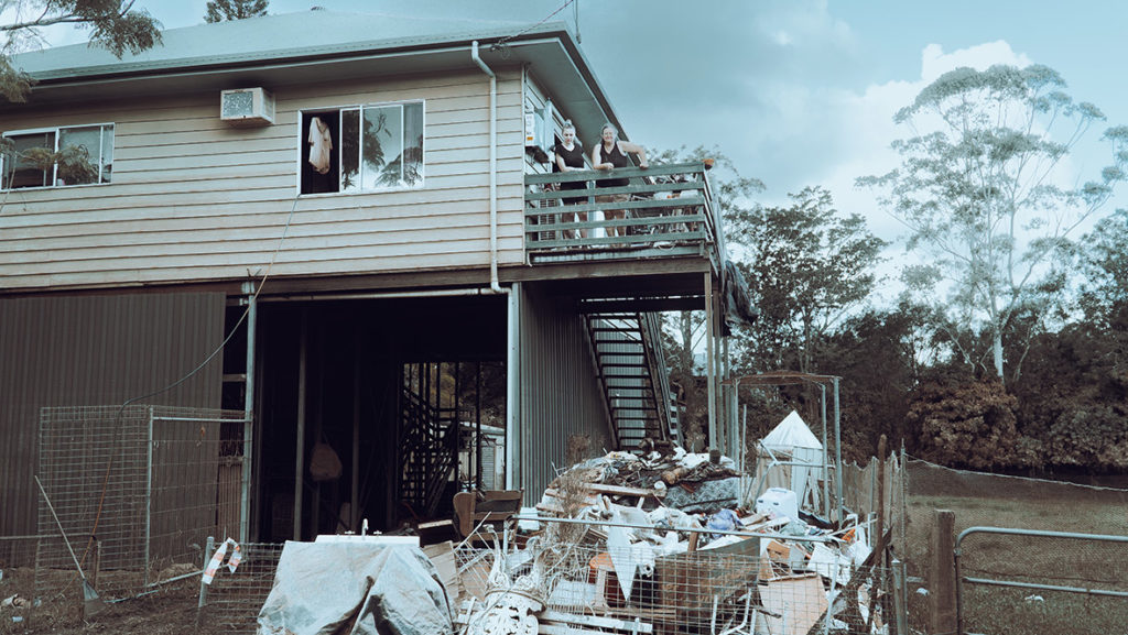 An image of 2 women standing on the balcony of a home destroyed by floods in Lismore.