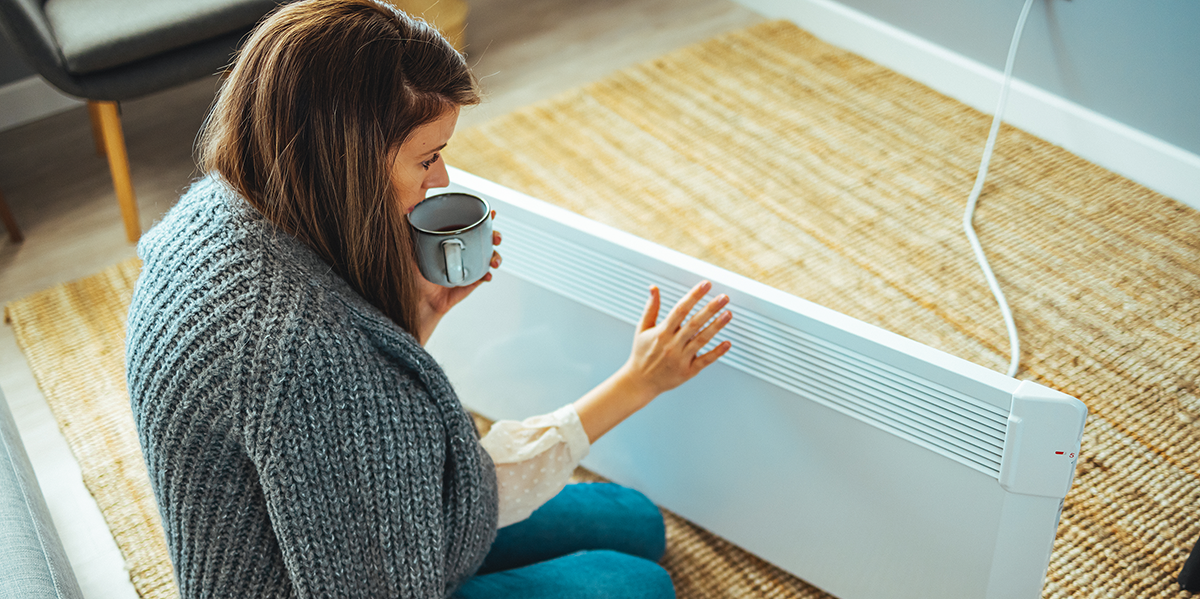 An image of a cold woman in front of a heater with a blanket and a hot tea.